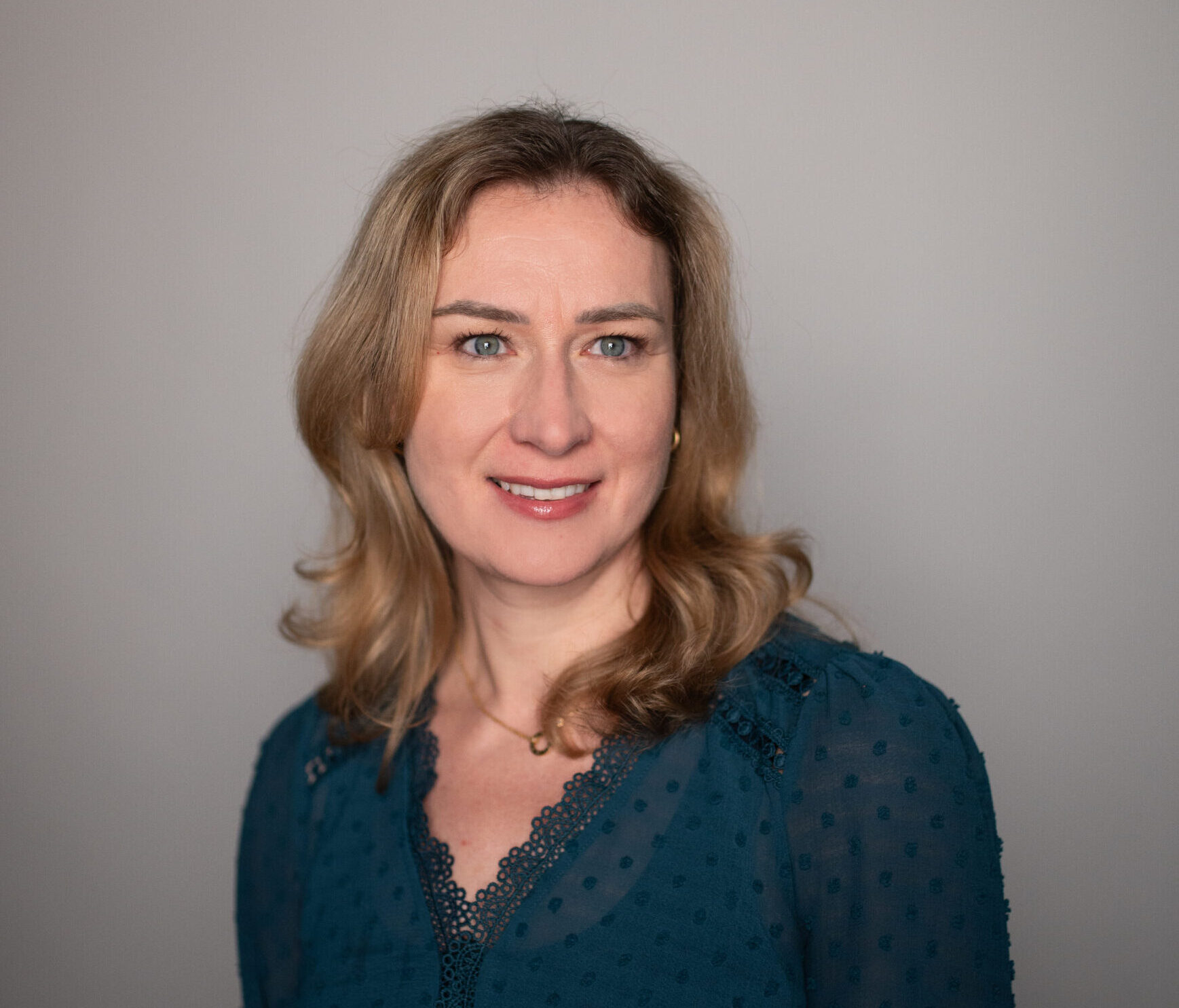 Portrait of a smiling woman with shoulder-length blond hair wearing a teal lace blouse against a neutral gray background.